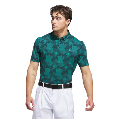 A man wearing an Adidas Mens Go-To Printed Polo Shirt in Pure Teal/Ivy, paired with white pants and a black belt, stands against a white background looking to his left.