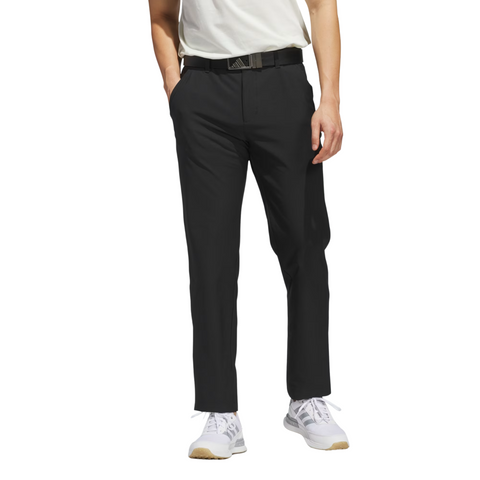A man models Adidas Ultimate365 Mens Golf Pants with a white t-shirt, black belt, and white sneakers, standing hands-in-pockets against a plain white background.