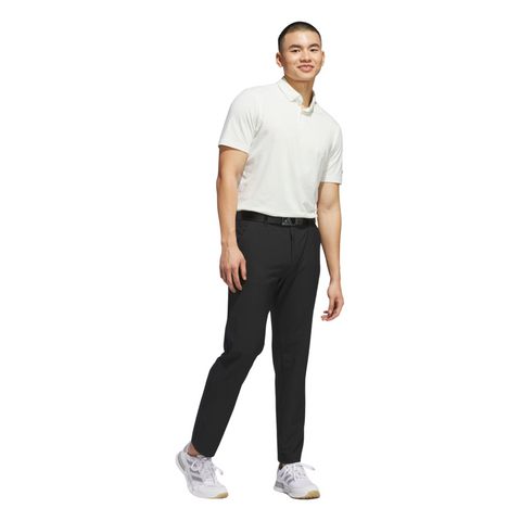 A man smiles, wearing a white short-sleeve polo shirt, black Adidas Ultimate365 Mens Golf Pants, a black belt, and white sneakers. He poses casually against a plain white background.