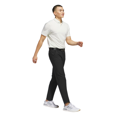 A man in a white polo shirt, Adidas Ultimate365 Mens Golf Pants by Adidas, and white sneakers walks confidently, looking ahead against a plain white background.