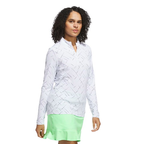 A woman with curly hair wears the Adidas Ultimate365 Printed Quarter-Zip Long-Sleeve Polo Shirt in white, paired with a light green ruffled skirt, standing against a plain white background.