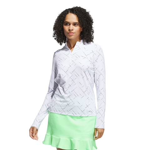 A woman with curly hair wears an Adidas Ultimate365 Printed Quarter-Zip Long-Sleeve Polo Shirt in white and a light green ruffled hem skirt, standing against a plain white background.