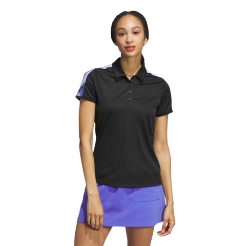 A woman with long braided hair and a neutral expression faces forward against a white background, wearing an Adidas Ultimate365 Womens Printed Polo in black and a purple skirt with a pocket.