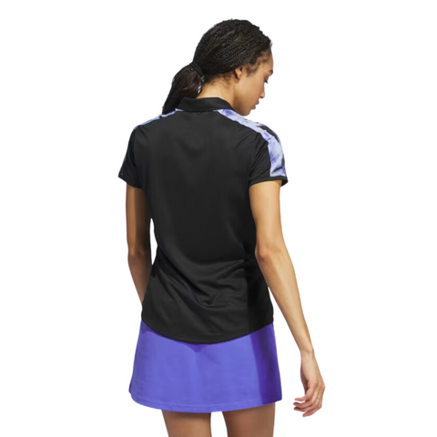 A woman with braided hair is seen from behind, wearing an Adidas Ultimate365 Womens Printed Polo in black with patterned shoulders and a purple skirt. The background is white.