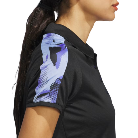 A woman in profile wears the Adidas Ultimate365 Womens Printed Polo by Adidas, with a black collar and a blue and white abstract shoulder pattern. Her long dark hair is styled in a ponytail.