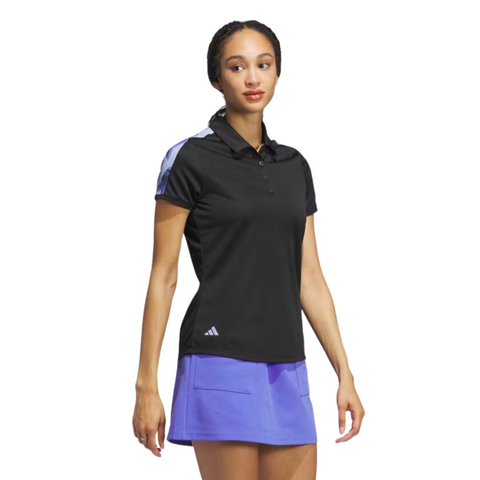 A woman faces forward against a white background, wearing an Adidas Ultimate365 Womens Printed Polo— a black, short-sleeve, moisture-wicking golf shirt with light purple accents— paired with a light purple skirt. Brand: Adidas.