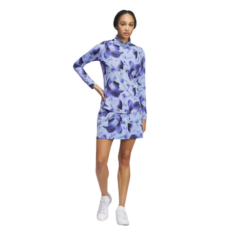 A woman wears an Adidas Ultimate365 Womens Skort with a blue and purple floral long-sleeve top and white sneakers, standing against a white background.