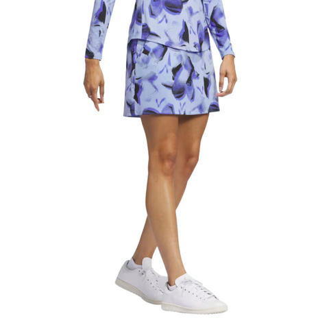A woman wears the Adidas Ultimate365 Womens Skort in a blue and purple floral print with a matching long-sleeve Adidas top and white sneakers, shown from shoulders to feet on a white background.
