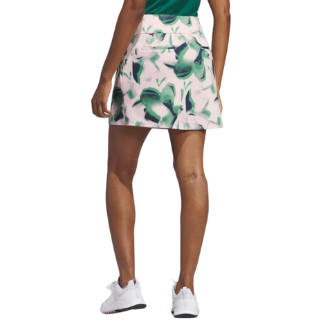 A person faces away from the camera, wearing a green top, white sneakers, and the Adidas Ultimate365 Womens Skort by Adidas in pink and green pattern, all set against a white background.