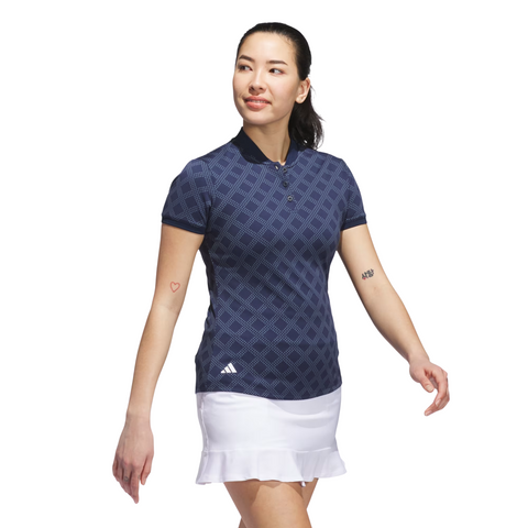A woman wearing an Adidas Womens Ultimate365 Jacquard Short Sleeve Polo Shirt in Collegiate Navy and a white skirt stands against a white background, showing small arm tattoos as she smiles and looks to the side.