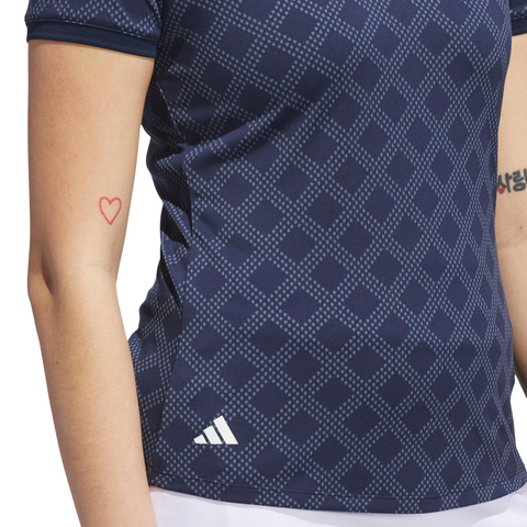 A person wears the Adidas Womens Ultimate365 Jacquard Short Sleeve Polo Shirt in Collegiate Navy, featuring a diamond pattern and side adidas logo. A simple red heart outline tattoo is visible on their left arm, with another tattoo partly seen on the right.