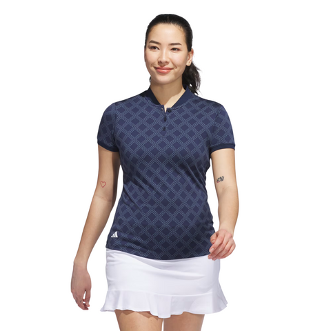 A woman wears an Adidas Womens Ultimate365 Jacquard Short Sleeve Polo Shirt in Collegiate Navy with a white skirt. She has a ponytail, small arm tattoos, and stands confidently against a plain white background.
