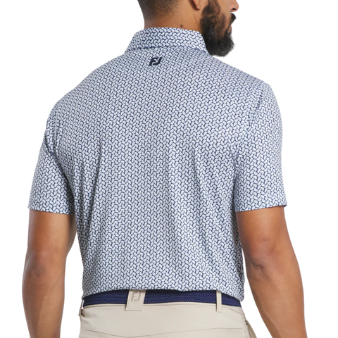 A man with a beard is seen from behind wearing a FootJoy Men’s Citrus Print Lisle Polo in White/Navy, made with ProDry fabric and featuring a small “FJ” logo beneath the collar, paired with beige pants.