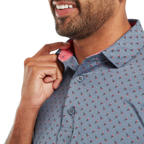 A man with a trimmed beard and mustache smiles while adjusting the collar of his blue FootJoy Men's Flower Bud Foulard Lisle Polo in Flint, featuring a stylish pattern, contrast pink inner collar, and moisture-wicking fabric.