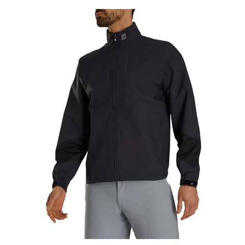 A man models the FootJoy Men’s HydroLite X Rain Jacket in black, featuring a stand-up collar, paired with light gray pants. One hand is at his side and the other extended, set against a plain white background.