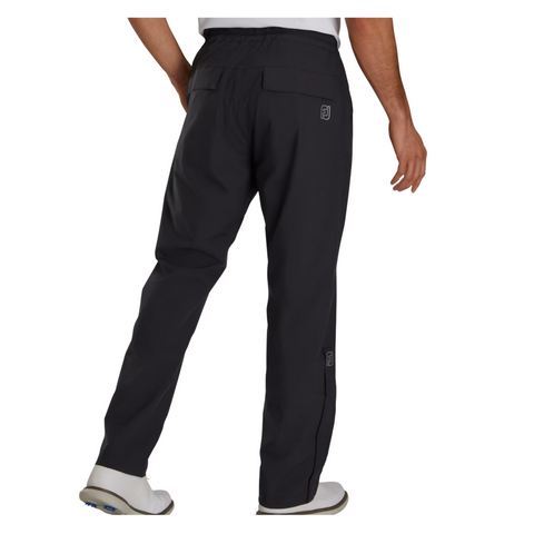 A person is seen from the back wearing black FootJoy Men’s HydroLite X Rain Pants and white shoes, with one arm slightly bent. The waterproof pants feature back pockets and a FootJoy logo near the right pocket.