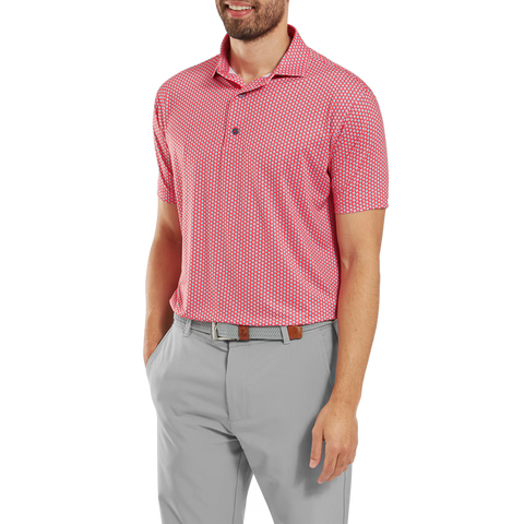 A man smiles with one hand in his pocket, wearing the FootJoy Mens Lisle Houndstooth Geo Print Polo in Hibiscus, tucked into light gray pants with a brown belt.