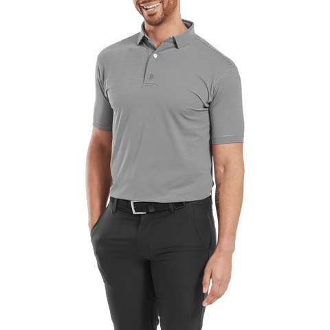 Smiling man wearing a FootJoy Mens Micro Feeder Stripe Lisle Polo in Black/Grey, tucked into black pants with a black belt, standing with one hand in his pocket.