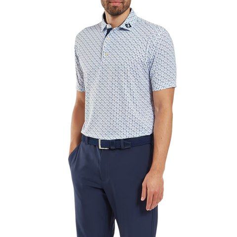 A man wears a FootJoy Mens Pique Solid Figure Print Lisle Polo in White/Navy, tucked into navy pants with a matching belt. His head is slightly turned and his left hand is in his pocket; the polo’s moisture-wicking fabric adds comfort.