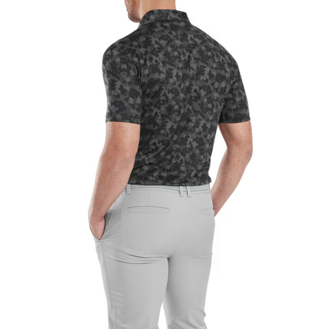 A man wearing a dark FootJoy Men's Pop Camo Lisle Polo in Black/Grey and light gray pants is shown from the back with his hands in his pockets against a white background.