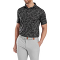 A man stands confidently with one hand in his pocket, wearing a FootJoy Men's Pop Camo Lisle Polo in black/grey, tucked into light gray pants and paired with a white woven belt.