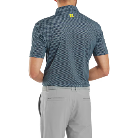 A man wearing a FootJoy Men’s Wiggle Print Lisle Polo in navy/white and light gray pants stands with his back to the camera. The polo features ProDry Performance Fabric and a small yellow logo at the upper back.