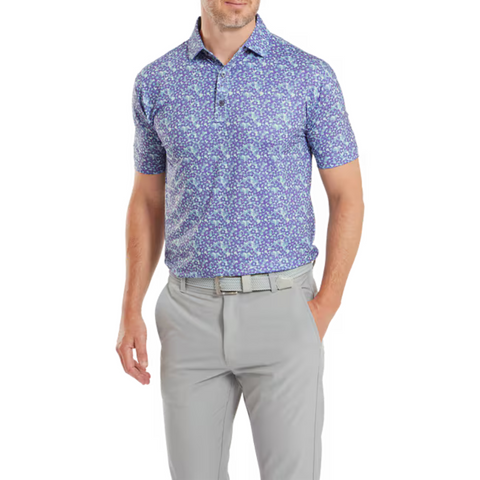A man wears the FootJoy Primrose Print Lisle Mens Polo, a short-sleeve blue and white golf polo by FootJoy, tucked into light gray pants with a matching belt. He stands against a white background with one hand in his pocket.