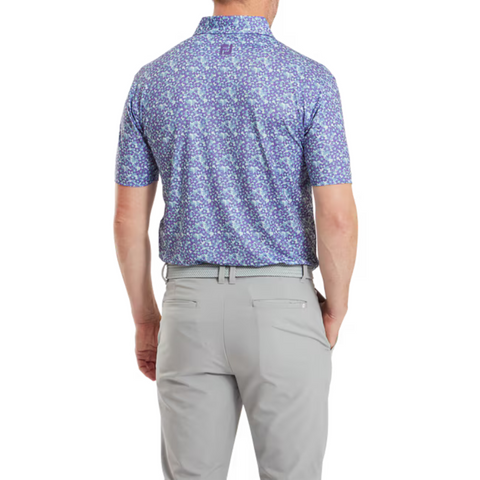A man faces away from the camera, wearing a FootJoy Primrose Print Lisle Mens Polo in purple and blue, neatly tucked into light gray pants with a belt. The background is plain white.