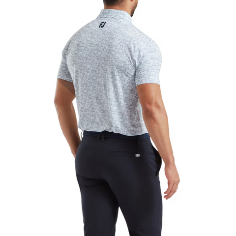 A man in a light gray patterned FootJoy Scottish Town Mens Print Polo and dark trousers stands facing away with hands in his pockets. The moisture-wicking polo has a small black logo near the collar.