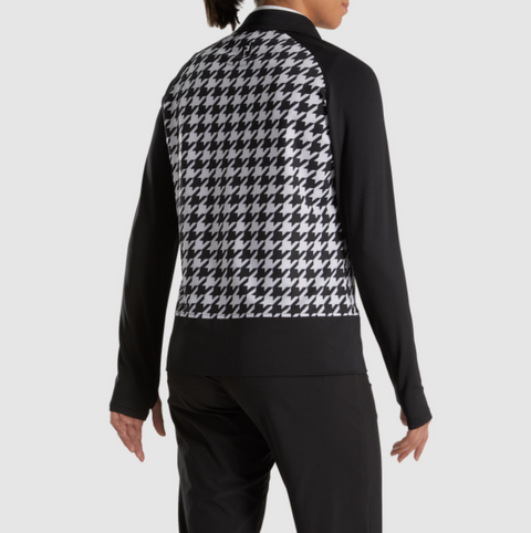 A person wearing the FootJoy Womens Full Zip Houndstooth Mid Layer Jacket in black/white stands facing away with arms relaxed, showcasing the white and black houndstooth pattern on the back against a plain, light background.