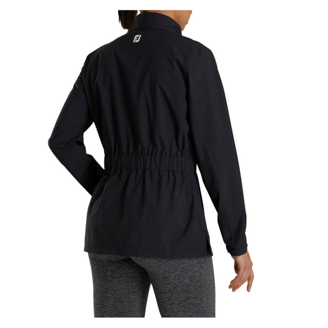 A person wearing gray leggings and a black FootJoy Women’s HydroLite Jacket with an elastic waistband is shown from the back; this waterproof golf jacket features a small white FootJoy logo near the collar.