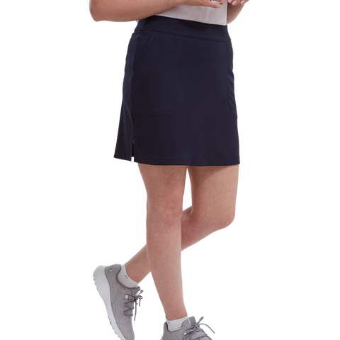 A woman shown from mid-torso down stands with one leg slightly bent, wearing a FootJoy Womens Interlock Stretch Skort in navy, light gray sneakers, and a white top.