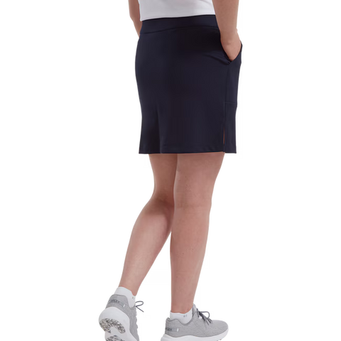 A person wearing a FootJoy Womens Interlock Stretch Skort in navy, a white shirt, and gray athletic shoes stands with hands in skort pockets, photographed from behind against a white background.