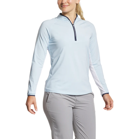 A woman stands smiling against a white background, wearing the FootJoy Women’s Long Sleeve Sun Protection Shirt in Skyway/Navy and light gray pants.