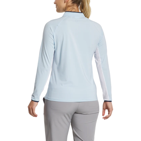 A woman with blonde hair in a ponytail is shown from the back, wearing FootJoy Women’s Long Sleeve Sun Protection Shirt in Skyway/Navy and gray pants against a white background.