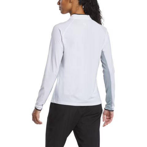 A woman with a ponytail, facing away, wears a FootJoy Women's Quarter-Zip Long Sleeve Sun Protection Shirt in white (UPF 50+) and black pants. Her left arm is bent slightly, and she stands against a plain white background.