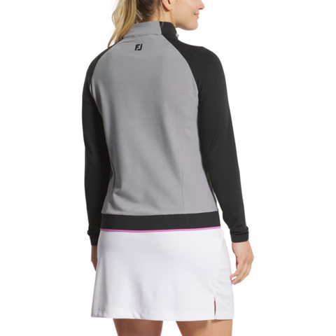 A woman faces away in a FootJoy Women's Quarter-Zip Midlayer Top in black/grey, featuring the FJ logo and four-way stretch fabric, paired with a white skirt with a small slit, against a plain background.