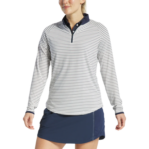 A woman wears a FootJoy Womens 1/4 Zip Long Sleeve Sun Protection Shirt—white with thin navy stripes and a navy collar—paired with a navy athletic skirt, standing against a plain white background.