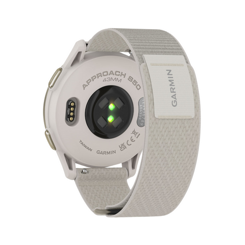 The back view of a Garmin Approach S50 GPS watch in beige shows the heart rate sensor glowing green, the charging port, and a fabric strap labeled GARMIN.