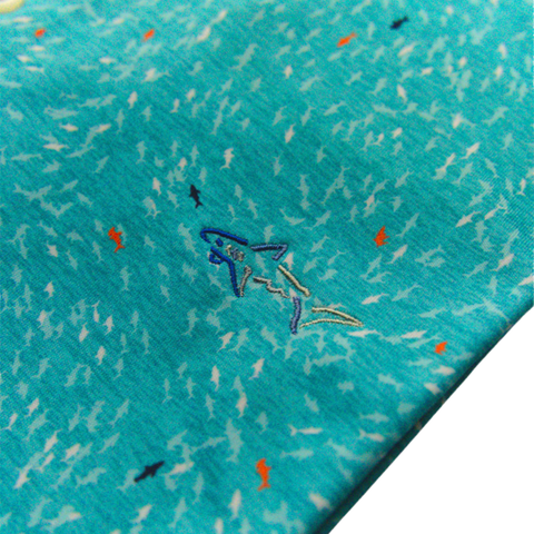 Close-up of Greg Norman Men’s ML75 Shark Frenzy Print Polo in Aquamarine, showing blue fabric with small white, black, and orange shark prints plus an embroidered blue shark—ideal detail for a stylish, moisture-wicking golf polo.