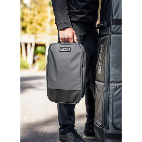 A person dressed in black holds an Ecco Ping Shoe Bag in gray, standing outdoors beside a larger matching golf bag with trees blurred in the background.