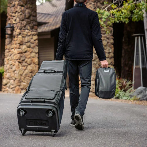 A person in black pants and a jacket walks on a paved path, pulling a large wheeled golf travel bag and carrying the Ecco Ping Shoe Bag, with stone buildings in the background.