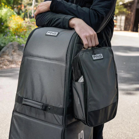 Outdoors, a person holds the handle of a large gray travel bag with an Ecco Ping Shoe Bag attached and resting against it. Trees and a pathway are visible in the background.