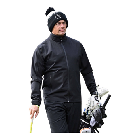 A man in a Proquip ProQuip Mens Aqualite Pro Rain Jacket, black pants, and a black beanie with a white pompom carries a golf bag and yellow club while walking outdoors.