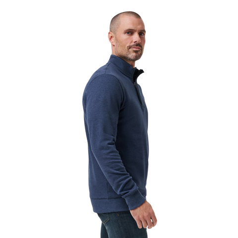 A man with a shaved head and short beard, wearing a Travis Mathew Cloud 1/4 Zip 2.0 Men’s Pullover in Heather Blue and jeans, stands sideways against a white background.