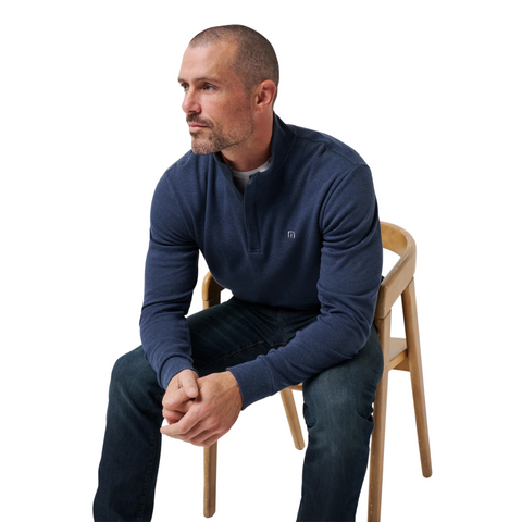 A man with short hair and a trimmed beard, wearing the TravisMathew Cloud 1/4 Zip 2.0 Men’s Pullover in Heather Blue and jeans, sits on a wooden chair looking to the side against a white background.