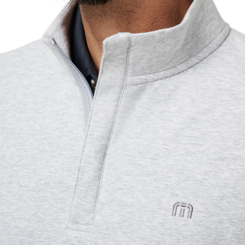 Close-up of a person wearing the Travis Mathew Cloud 1/4 Zip 2.0 Men’s Pullover in Heather Light Grey, featuring a lightweight, ultra-soft fabric, standing collar, partially open zipper revealing a dark shirt, and an embroidered chest logo.