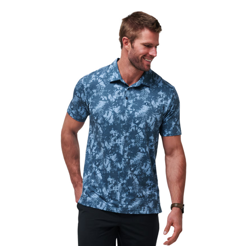 A man in a blue Travis Mathew Featherweight Please Mens Polo with a light blue floral pattern and black pants smiles while looking left against a white background.