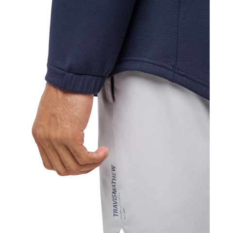 A close-up of a person’s left hand near their hip, wearing the Travis Mathew Cloud 1/4 Zip 2.0 Men’s Pullover in Heather Blue and light gray shorts with TRAVISMATHEW printed vertically on the shorts.