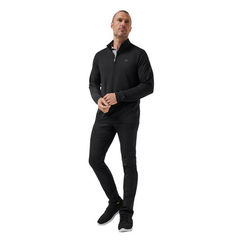 A man stands against a white background, wearing the TravisMathew Heater Pro 1/4 Zip Men’s Pullover in black by Travis Mathew, paired with black pants and black sneakers with white soles. He gazes slightly to the side with his hands loosely clasped.